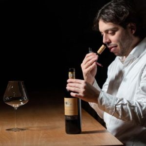 Wine tasting con sommelier che annusa il tappo di una bottiglia, calice vuoto su tavolo di legno.