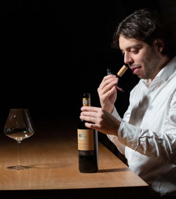 Wine tasting con sommelier che annusa il tappo di una bottiglia, calice vuoto su tavolo di legno.